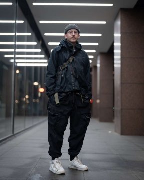 Ein neben einem futuristischen Gebäude in kompletten Techwear Outfit bestehend aus einer ACRONYM Techwear Jacke, einer ACRONYM Techwear Tasche, einer ACRONYM P38-E Hose und ACRONYM x Nike Lunar Force Techwear Sneaker