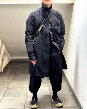 Ein Mann in einem futuristischen Techwear Outfit bestehend aus ACRONYM J1L-GT Techwear Mantel, ACRONYM J97-M Techwear Jacke, ACRONYM P30A-E Techwear Hose und Nike Mountain Fly GTX Techwear Schuhe