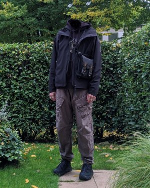 Eine Person in stehend in einem Garten in einem Techwear Outfit mit Adidas MyShelter Techwear Jacke, Enfin Levße Aurrean Techwear Hose und Hoka Tor Low Techwear Schuhe und ACRONYM 3A-2 Tasche